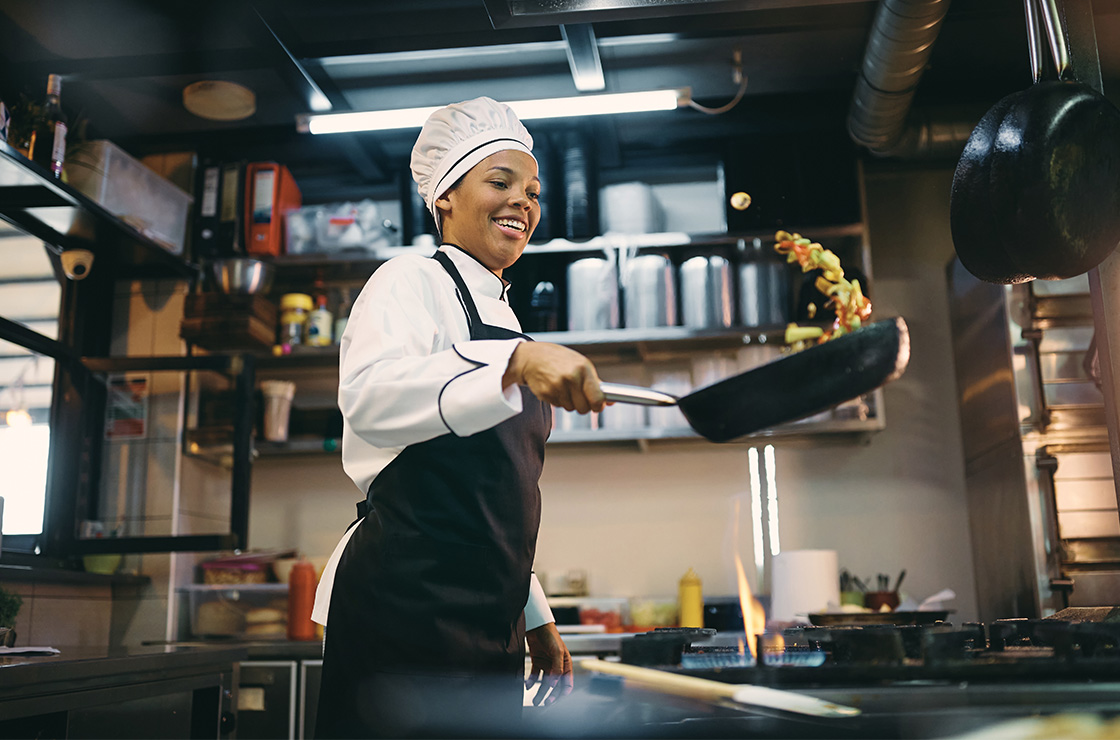 Happy black female chef preparing food in frying pan at restaurant kitchen